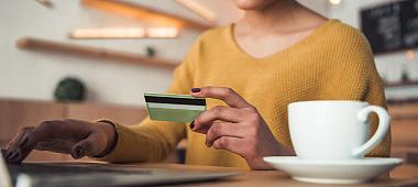 Cropped image of beautiful Afro American girl drinking coffee and holding a credit card while paying online with a laptop in cafe